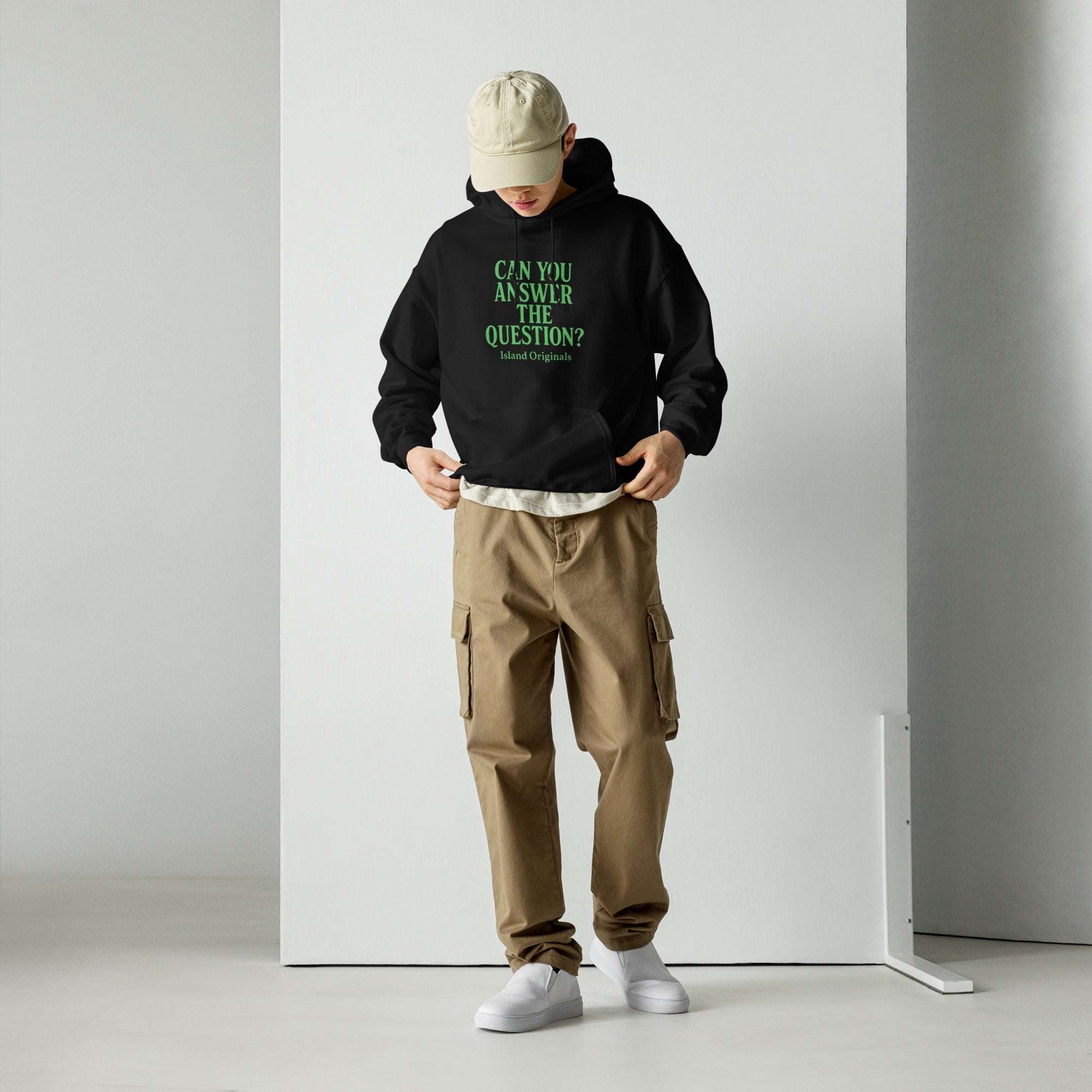 Person wearing a black hoodie with text and beige cap, standing against a white wall.