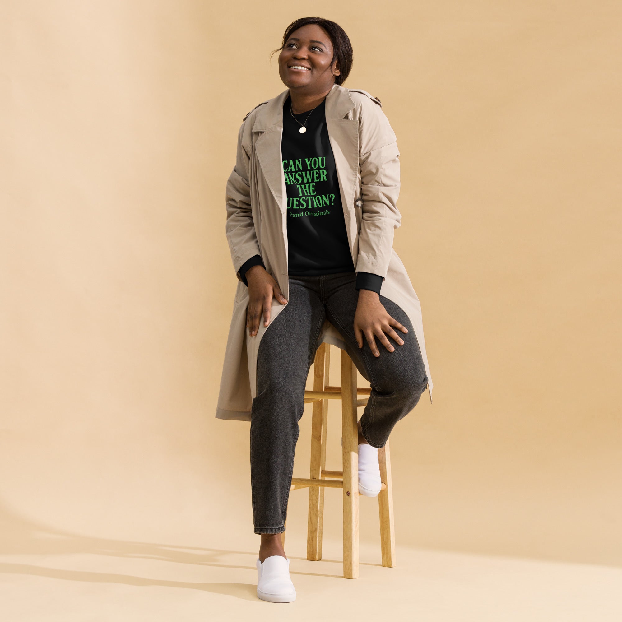 Woman sitting on a stool wearing a trench coat and black shirt with text, against a beige background